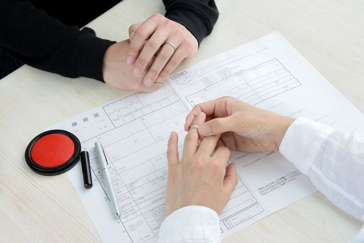 離婚届を挟んで相対して座るカップルの1人が、結婚指輪を外している