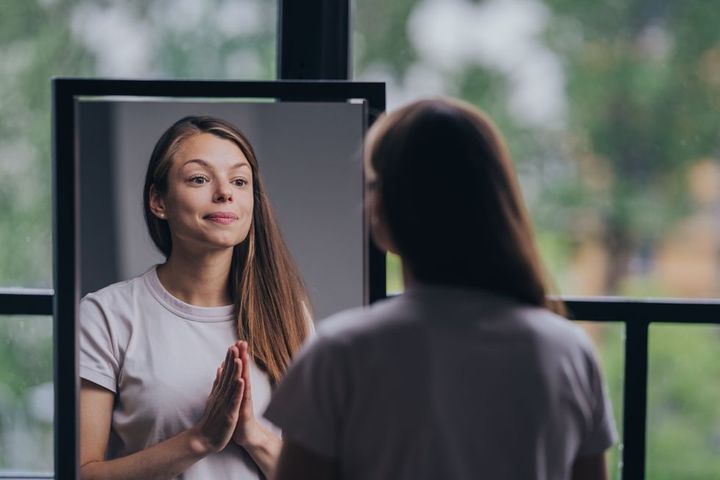 reflective young woman in a casual t shirt practicing self affirmation in front of a mirror in a well lit room