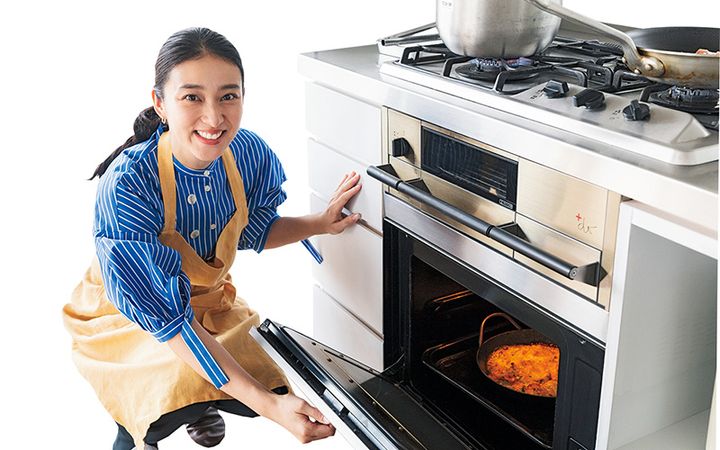 キッチンでオーブンの扉を開けるエプロン姿の武井咲さん