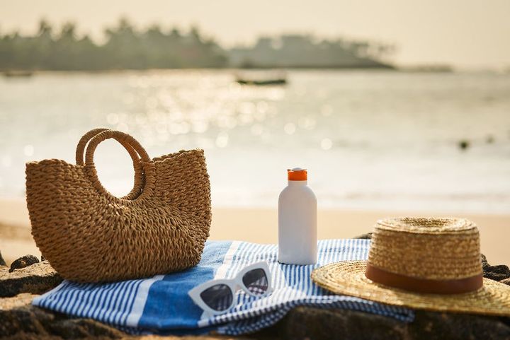 Protective sunblock essential for tropical climate. Beach scene with sunscreen bottle, straw hat on blue towel. UV protection gear, sunglass on sunny ocean shore. Holiday, skin care, leisure concept.