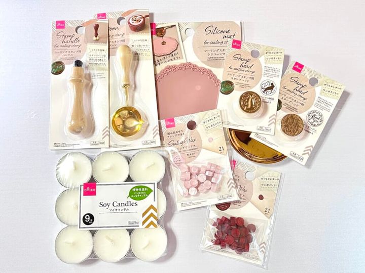ダイソーのハンドメイド用品「シーリングスタンプ用 ハンドル」「シーリングスタンプ用 スタンプヘッド」「シーリングスタンプ用 スプーン」「シーリングスタンプ用 シリコーンマット」「シーリングワックス 粒タイプ」「ソイキャンドル 9個入り」を机に並べて紹介する画像