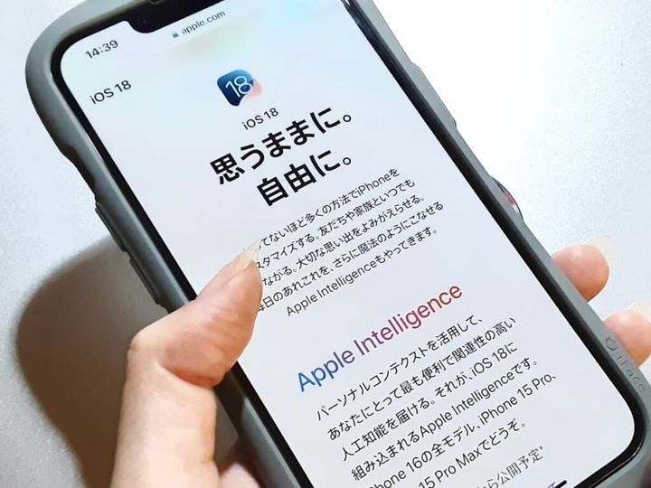 iPhoneを手に持ち、Apple公式サイト「iOS 18正式リリースページ」を紹介する画像