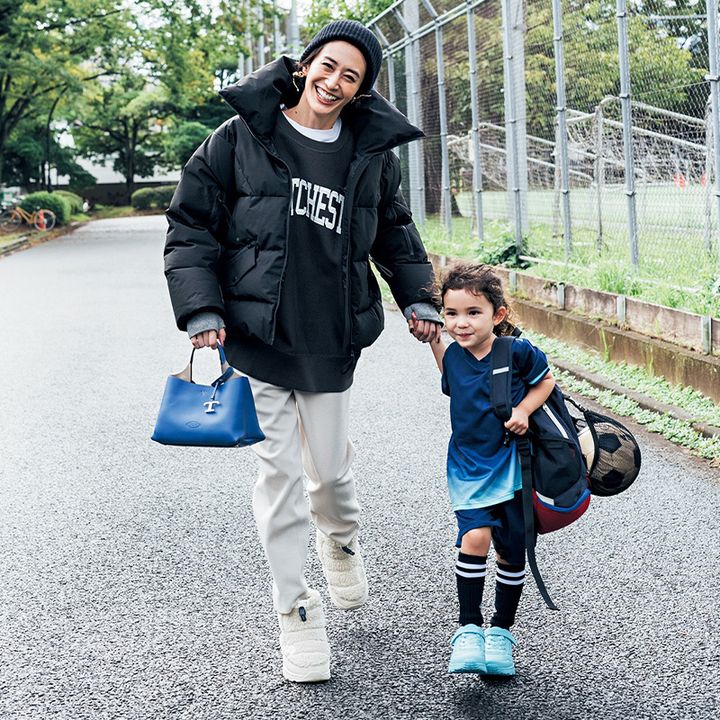 黒のスタンド襟のダウンコートに黒のロゴスウェットと白のスウェットパンツを着たママがサッカーユニフォームを着た子どもの手を引いている