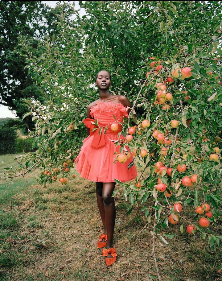 Model wearing pink, strapless, ruffle mini dress and pointed toe shoes with bow standing next to apple tree