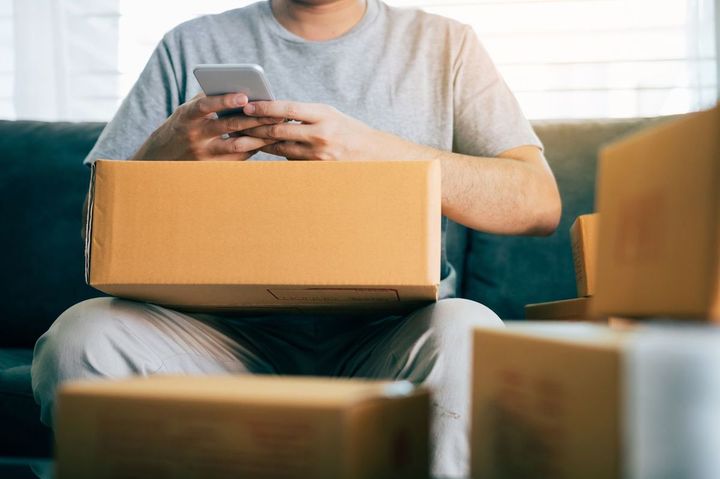 段ボールをひざにのせ、ソファーに座ってスマホを使用している男性