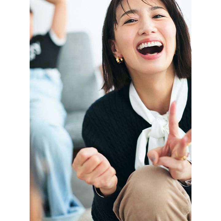 白のボウタイブラウスに紺のカーディガンを着て笑顔でピースする東原亜希さん