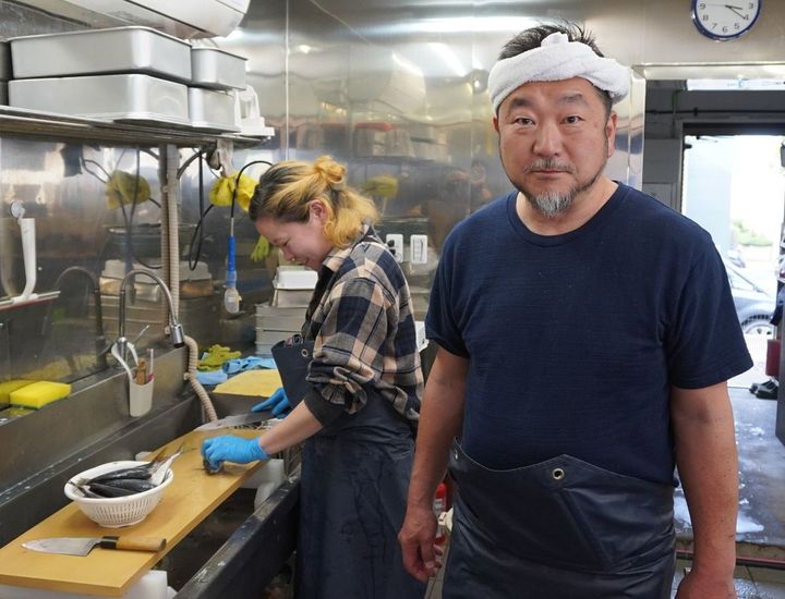 魚さばきの初心者にはアジがおすすめと説く上田勝彦さん。後方でアジをさばきながら笑っているのはサカナヤマルカマ店長の和田あかねさん。