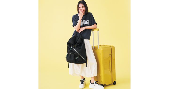 ロゴTシャツと白スカートに大容量黒リュックとホーガンの白スニーカーを合わせてリモワの黄色のスーツケースに肘をかけた岡本あずささん