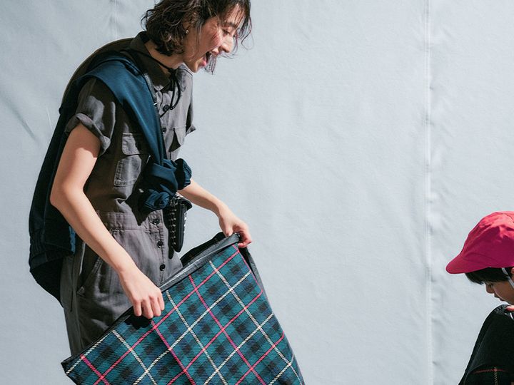 グレーの半袖オールインワンにジャケットを襷掛けしてチェックのレジャーシートを持つママ