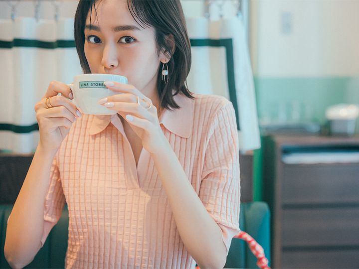 ピンクのポロニットにパールのついた大ぶりピアスと大ぶりリングをつけてラテを飲む桐谷美玲さん