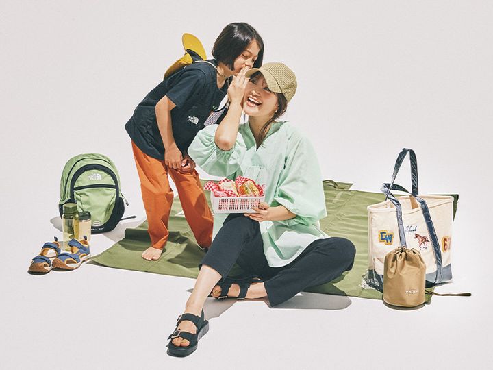 グリーンのボリューム袖ブラウスとスティックパンツにサンダルやキャップを合わせてシートに座って子どもと話すママ