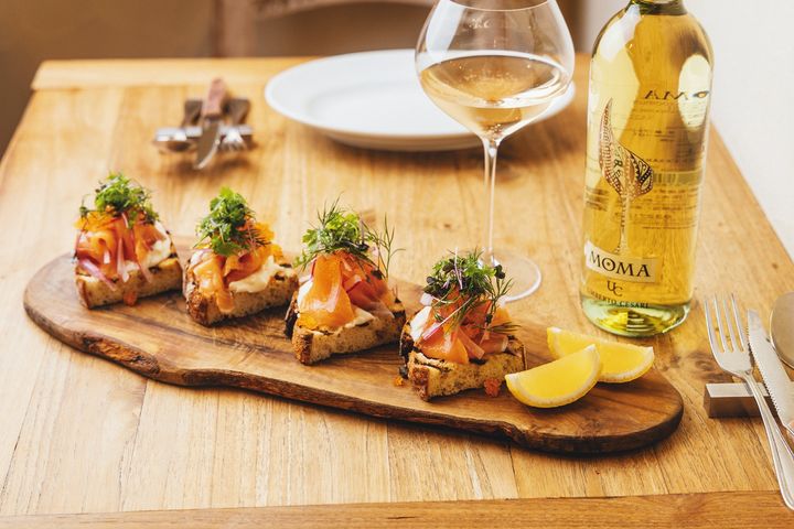 サーモンと赤玉ねぎのスライスをマリネした自家製スモークサーモンとストラッチャテッラのブルスケッタ1枚￥500（注文は2枚～。写真は4枚）。モマ・ビアンコ 2023（グラス）￥1,200。