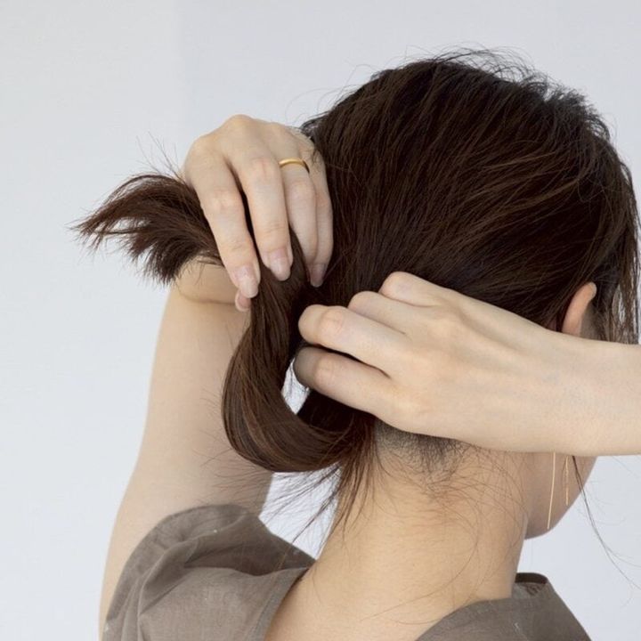 【HOW TO】1．衿足に向かって手ぐしでざっくりとまとめたら、ゴムでひとつに縛ります。次に根もとを押さえたまま、毛先側から毛束をクルンと折りたたむように持ち上げます。