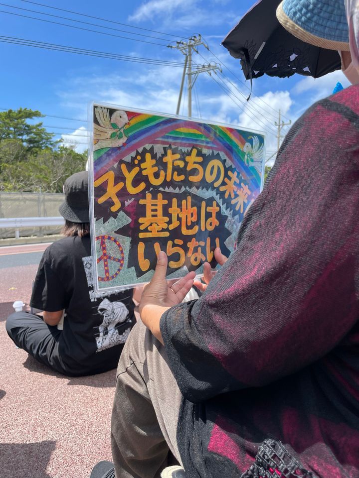 キャンプ・シュワブのゲート前での抗議の様子。前列に座っていた方が、「子どもたちの未来に基地はいらない」と書かれたプラカードを掲げていた。