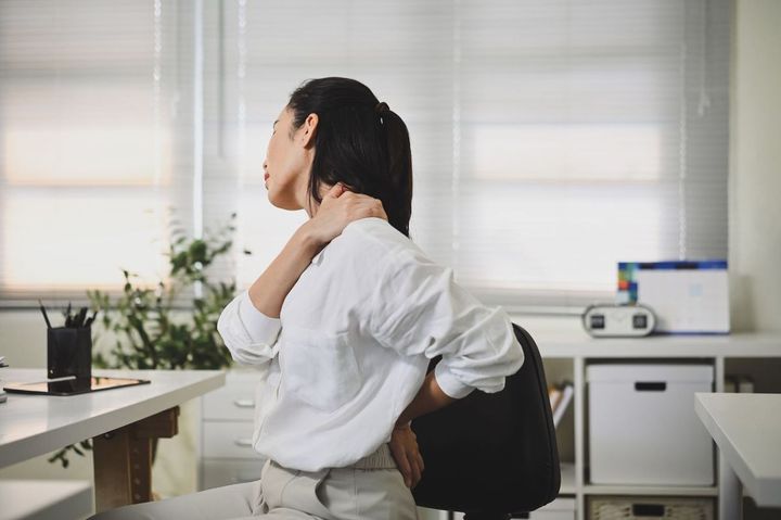 オフィスの自席で肩の張りを感じて、手を当てている女性