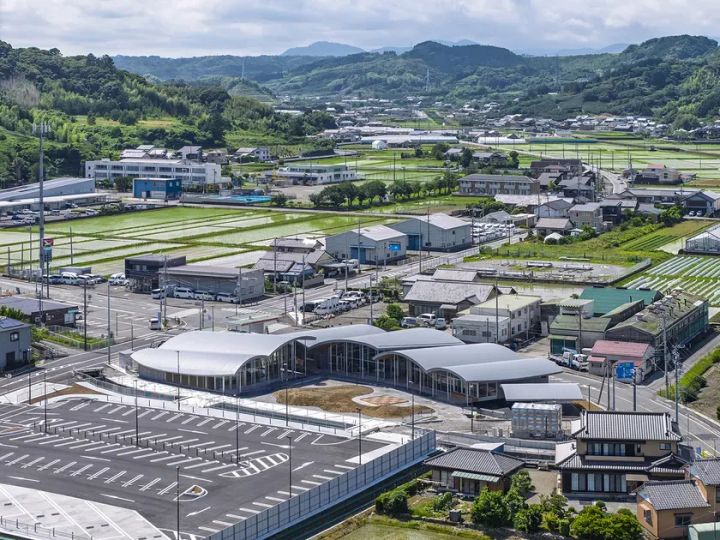 山の麓にある街並みの風景写真。手前には駐車場と特徴的なデザインの建物があり、奥には田んぼや住宅地が広がっている。全体的に緑豊かな自然と調和した美しい景観である。