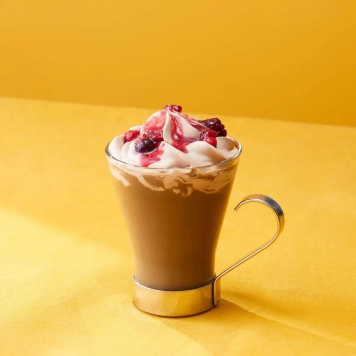 ホイップクリームとベリーソースがトッピングされたコーヒーが入ったガラスのマグカップ。背景は黄色。