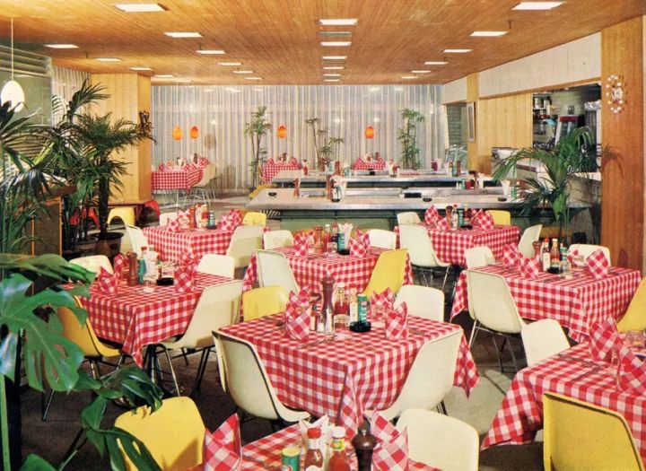 レトロな雰囲気のレストランのインテリア。テーブルには赤いギンガムチェックのテーブルクロスがかけられ、白い椅子が並んでいます。観葉植物が配置され、照明が天井から吊り下げられています。