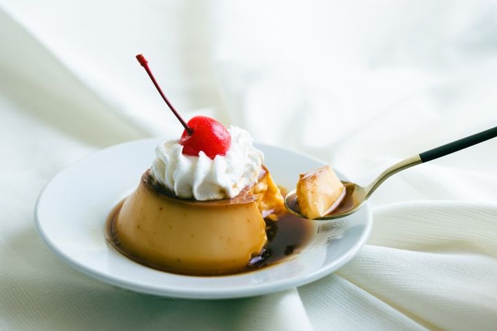 白い浅皿にクラシックプリン ホイップクリームとてっぺんにさくらんぼ プリンをスプーンですくう様子