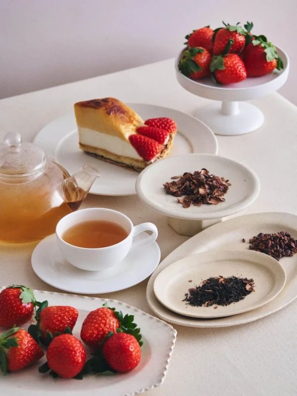 テーブルの上に、イチゴ、ケーキ、お茶、チョコレート、紅茶が置かれた写真です。イチゴは、ケーキの上にトッピングとして、また、皿の上に盛り付けられています。ケーキは、クリームとスポンジの層があり、焼き目がついています。お茶は、ティーポットとカップに注がれており、チョコレートと紅茶は、それぞれ別の皿に盛られています。