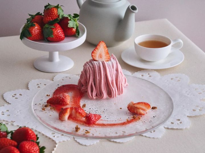 いちごのモンブランケーキと紅茶のティーセットの写真。モンブランケーキはピンク色のクリームで覆われ、上にいちごのスライスが乗っている。周りにはカットされたいちご、いちごソース、ティーカップ、ティーポットが配置されている。