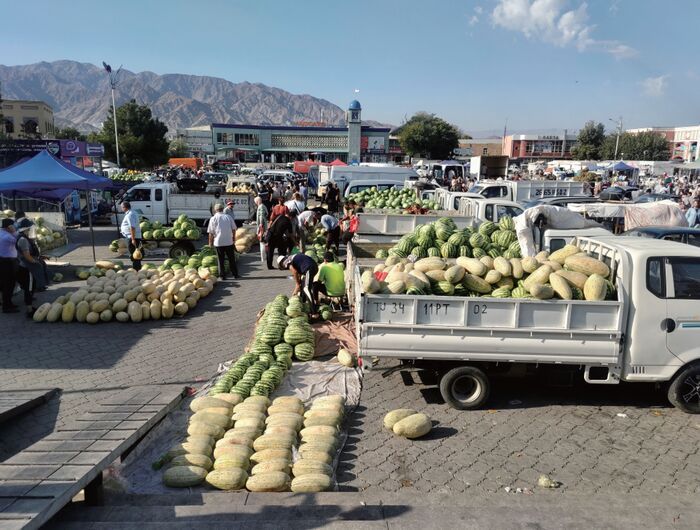 屋外の市場の様子。トラックの荷台や地面に大量のスイカやメロンが並べられ、人々が売買や積み下ろしをしている。背景には山並みが広がっている。