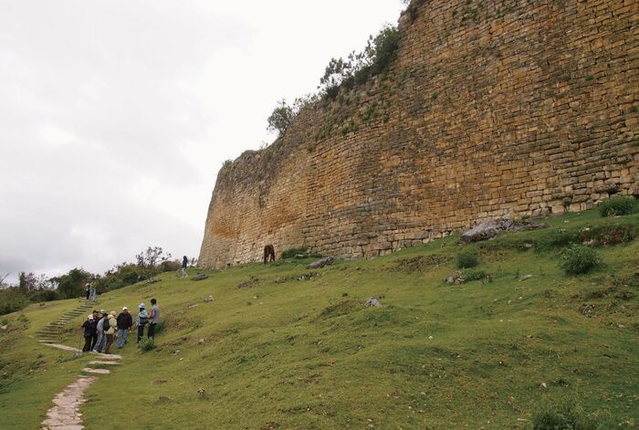 ペルー北部のチャチャポヤス。草原の斜面にそびえ立つ巨大な石造りの城壁と、その下を歩く人々。自然と遺跡が一体になった風景。