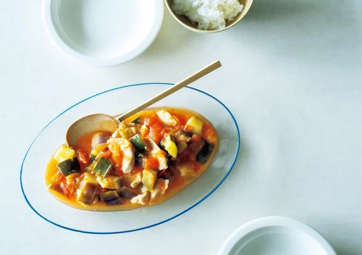 白いテーブルの上に、ラタトゥイユのような野菜と鶏肉の煮込み料理が盛られたガラスの皿、ご飯、白いお皿が置かれた写真です。