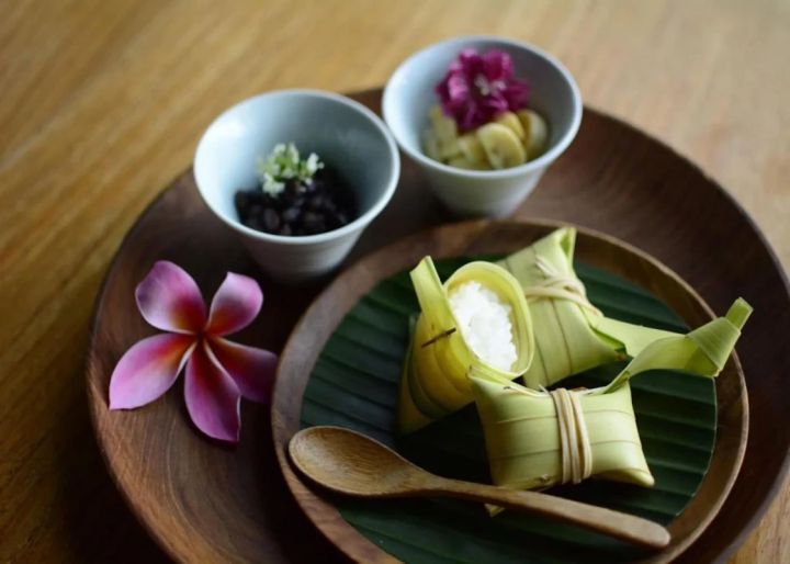 木製の皿の上に、ココナッツの葉で包まれたもち米、黒豆、バナナ、花が添えられた料理の写真です。