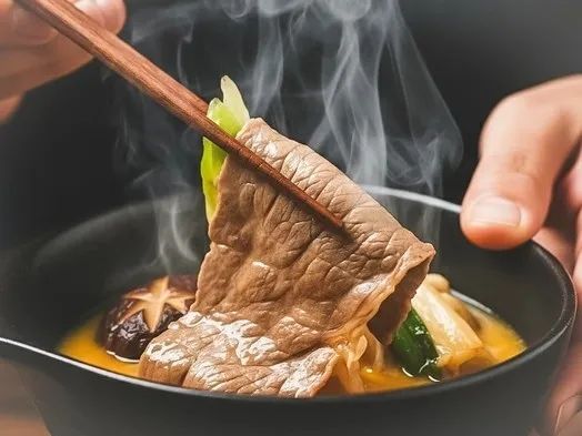 湯気の立つ鍋から持ち上げた牛肉が印象的なすき焼きのイメージ