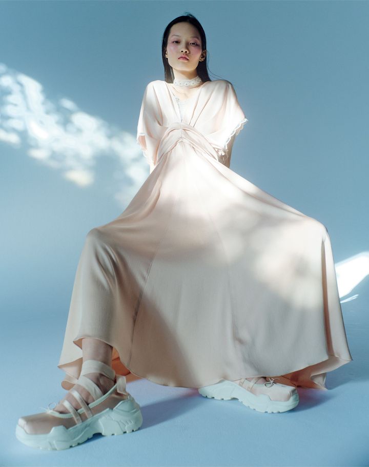 ユニフォームをフレッシュに再解釈 <br /> 青木明子はセントラル・セント・マーチンズなどでファッションを学んだ後、2014年に自身のブランドを設立。一貫してユニフォームをモチーフとしている。26年春夏はユニフォームを現実のメタファーと捉え、「内」と「外」の装いを融合。センターにレースをあしらったサテンのワンピースの足もとは、あえてスニーカーソールのシューズを。トゥシューズを思わせるディテールが、モダンかつフェミニンなムードを後押しする。ワンピース ￥86,900 シューズ（取り外し可能なリボンストラップ付き） ￥34,100 チョーカー 参考商品╱すべてAKIKOAOKI（アキコアオキ）