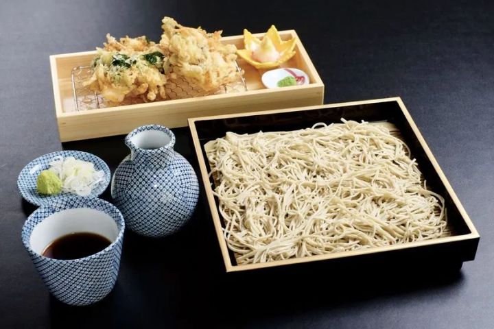 白っぽい手打ちそばをせいろに盛り、天ぷらと薬味を添えた和食膳の料理写真。網を敷いた盆には葉物やかき揚げ風の天ぷらが並び、染付の器にはそばつゆや薬味が用意されている。黒い卓上で器の藍色が映え、名物の手打ち蕎麦と揚げ物の質感を丁寧に伝える構成である。