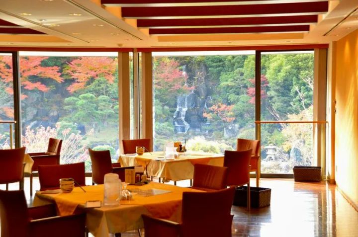 大きな窓越しに紅葉した日本庭園を望むレストラン空間の写真。店内には赤系の椅子と円卓が並び、天井や床には木の質感を生かした落ち着いた内装が広がっている。窓外には滝や岩組み、松が見え、景観と食事を一体で楽しめる上質なクラブハウスレストランの雰囲気を伝える構図である。