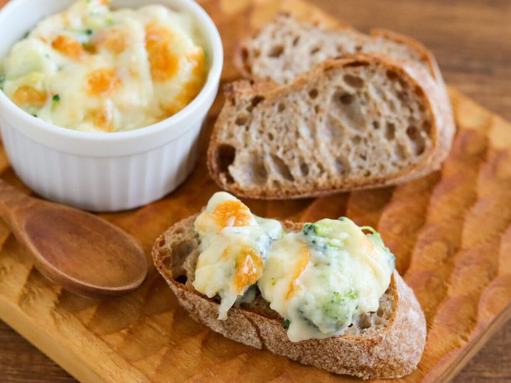 木のまな板の上にブロッコリーグラタンの皿とグラタンをのせたバゲット一切れが並んでいる様子