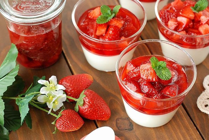 グラスに盛り付けたパンナコッタにいちごソースとミントをのせた完成品と保存瓶、手前にいちごとイチゴの花を添えた木製テーブルのスタイリング写真