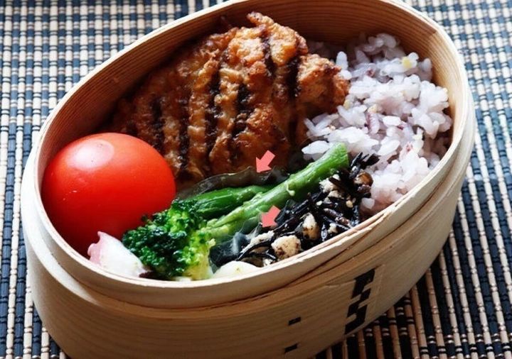 曲げわっぱに赤飯・唐揚げ・ブロッコリー・トマトなどを詰めた弁当に透明おかずカップを使った使用例、赤い矢印でカップの位置を示す