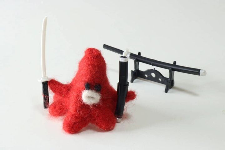 赤いぬいぐるみに白い長刀を持たせ刀掛けに飾った刀を並べた写真