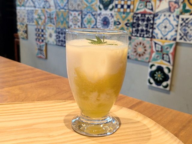 グラスの底に黄色いパイナップルソース、上にアーモンドミルクのフォームが重なりローズマリーが添えられたドリンク