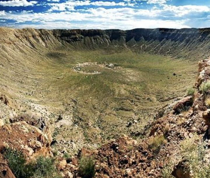 Wide view of a large circular crater with a green grassy floor and rugged rocky rim under a blue sky.