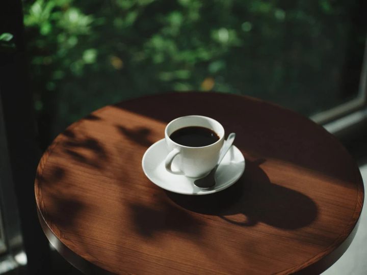 木製のテーブルに置かれたコーヒーカップの写真。カップは白く、コーヒーが注がれている。背景にはぼやけた緑の木々が見える。