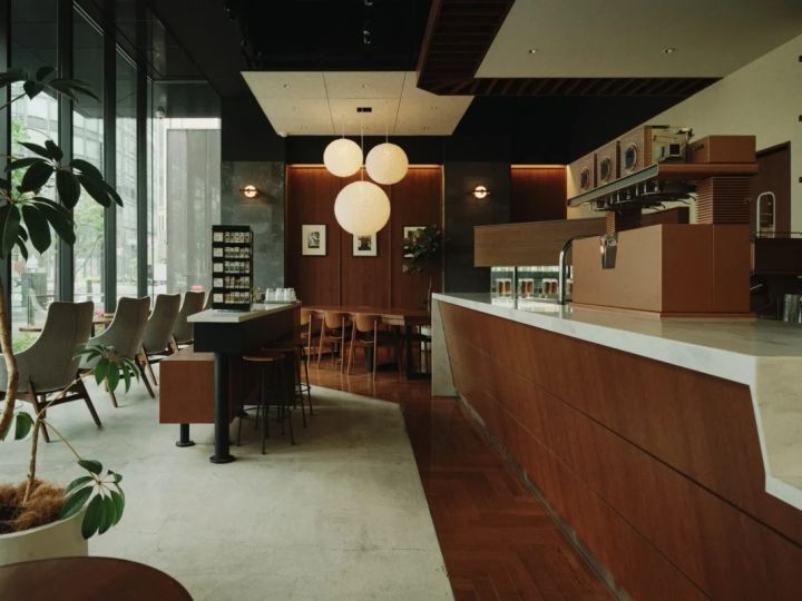 モダンな雰囲気のカフェの内観。木材とコンクリート、そして大きな窓から差し込む自然光が印象的。洗練されたデザインの照明器具や家具が配置され、落ち着いた空間を演出している。