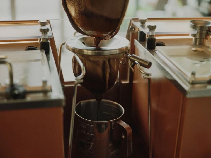 コーヒーを抽出している様子を捉えた写真です。コーヒーメーカーからコーヒーが抽出され、金属製のカップに落ちています。コーヒーメーカーとカップは、木製のフレームに囲まれた、透明なガラス面の上に置かれています。