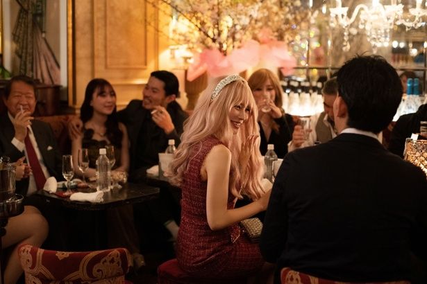 ネオンきらめく店内でキャバ嬢として完璧な笑顔で接客する麻由 [c]2026「マジカル・シークレット・ツアー」製作委員会 