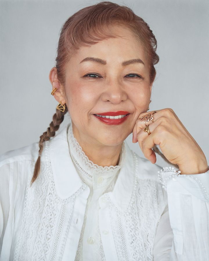 Smiling older woman with pale pink hair in a braid, wearing a white lace blouse and gold earrings and rings, resting her chin on her hand.