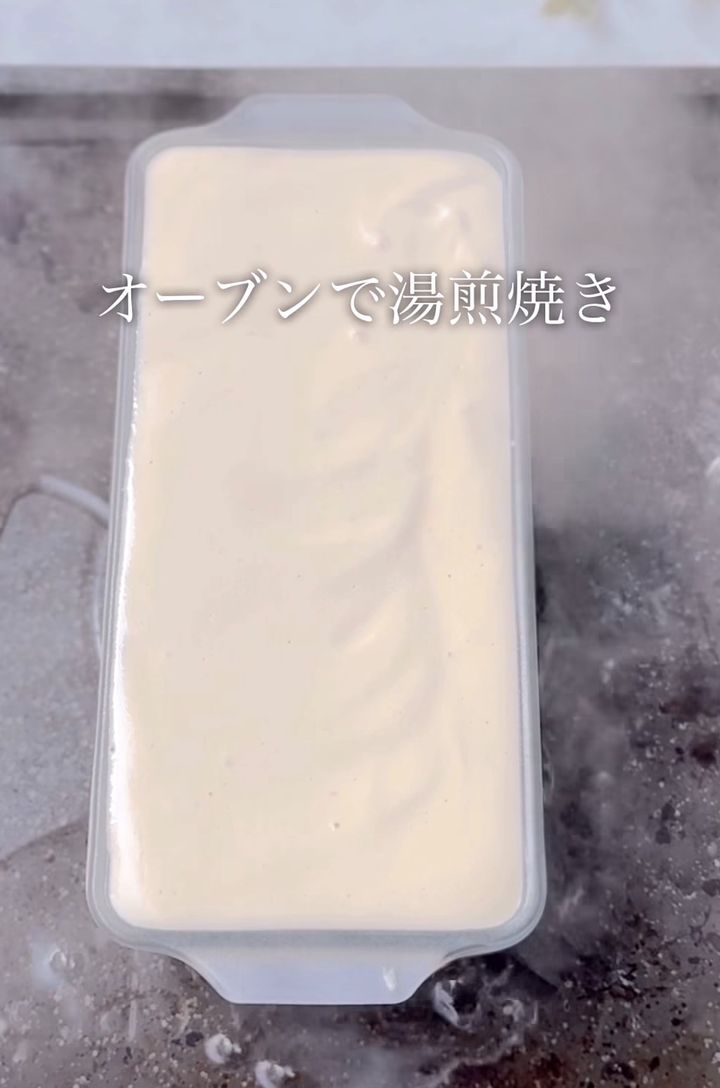 Pale creamy batter in a rectangular plastic dish with a translucent lid; overlay text reads 'オーブンで湯煎焼き' (baked in a hot water bath).