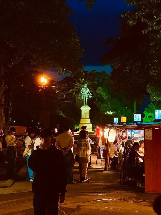 夜間の甲突川左岸緑地で提灯や屋台の明かりが灯る中、大久保利通銅像をバックに来場者が屋台に集まり飲食を楽しむ実際の開催風景