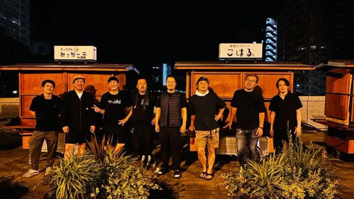 夜の甲突川沿いに並ぶリヤカー屋台「みっちー亭in鹿島」「串焼きスタンドこはる」の前で、黒Tシャツ姿の男性スタッフ8名が横一列に並んで記念撮影している集合写真