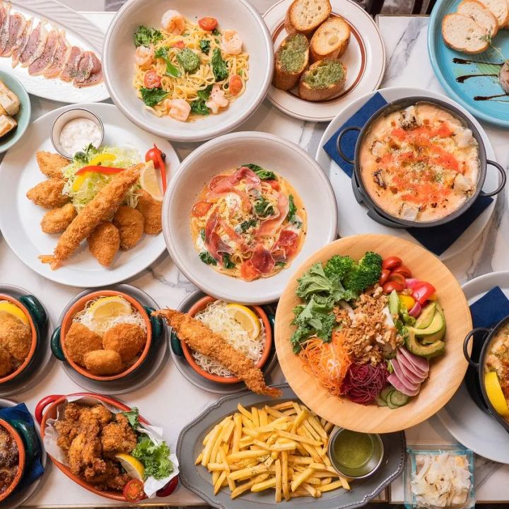 マルブルテーブルに並んだ多品目の料理フラットレイ。エビフライ・海老のパスタ・生ハムパスタ・フリット・フライドポテト・サラダボウル・グラタンなど10皿以上が俯瞰で写る