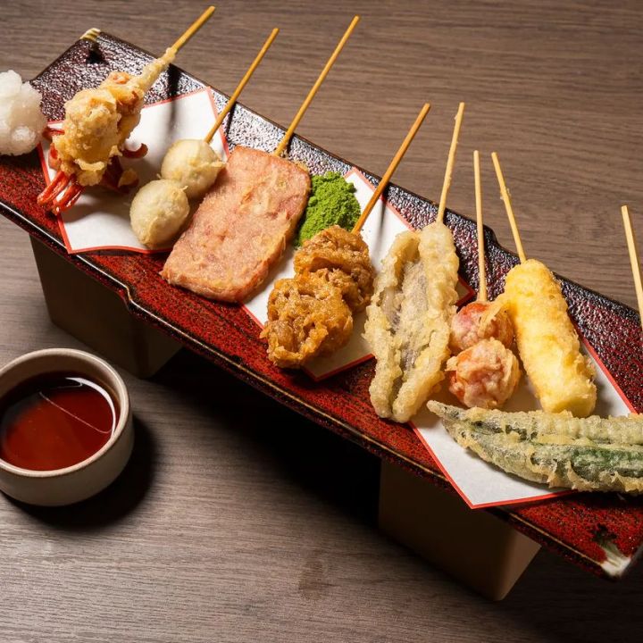 漆塗り長皿に並んだ串揚げ・天ぷら盛り合わせ。海老・ちくわ・スパム・野菜など約10本が竹串付きで整列し、左手にトマトソース小皿を添えた構成