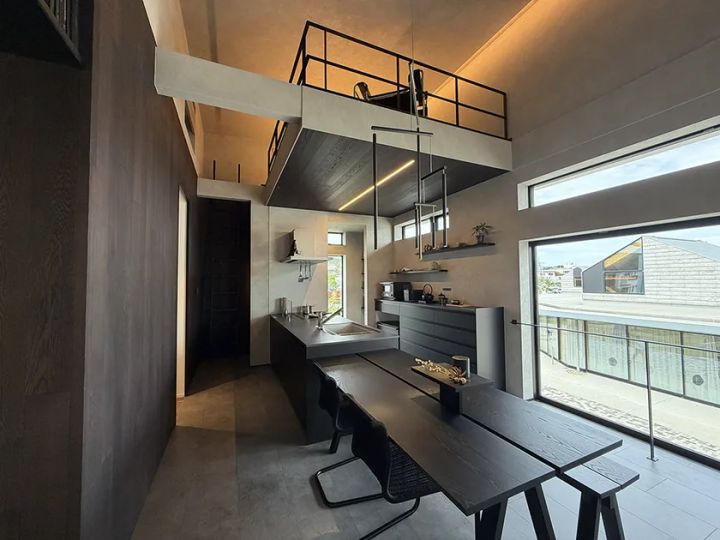 モデルハウス内部のダイニングキッチン実写。黒系アイランドキッチンとダイニングテーブル、鉄骨手すりのロフトスペースを上部に持つ吹き抜け空間